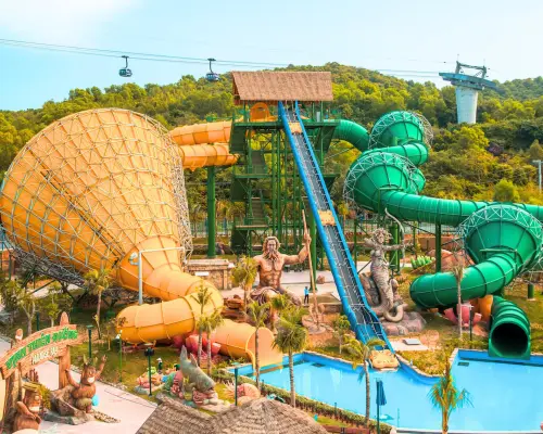 Panoramic view of Aquatopia Water Park in Phu Quoc, featuring diverse large water slides and aquatic facilities nestled in a lush green valley, with cable cars passing in the distance.