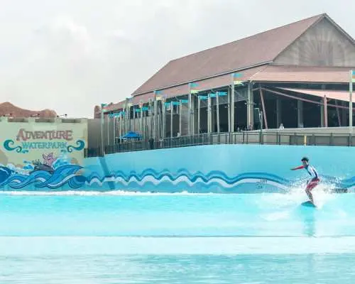 A visitor skillfully surfing on artificial waves in the large wave pool at Desaru Coast Adventure Waterpark, with the park's main building and sign visible in the background.