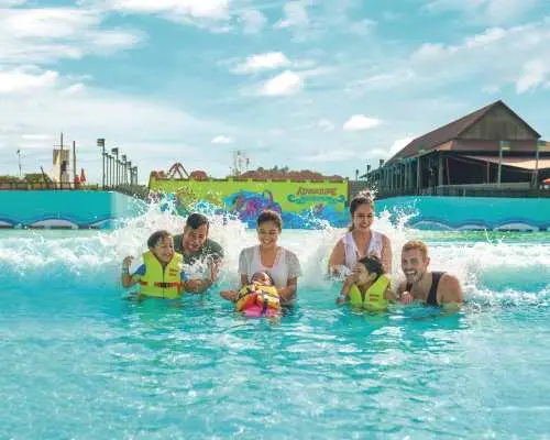 A happy family, including parents and two children in life jackets, joyfully playing in the gentle waves of the wave pool at Desaru Coast Adventure Waterpark.