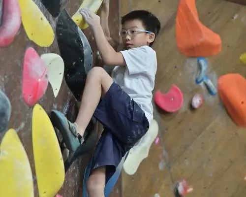 戴眼鏡的小男孩於JUST CLIMB室內抱石牆上積極攀爬，享受挑戰，體驗新手抱石課程的樂趣。