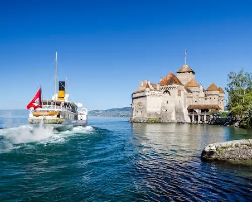 A paddle steamer flying the Swiss flag departs from Chillon Castle on Lake Geneva, creating ripples under a clear blue sky, combining historic architecture with scenic lake travel.
