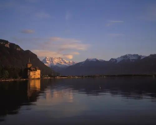 Panoramic view of Chillon Castle from across Lake Geneva at dusk, with its reflection on the calm water and snow-capped Swiss mountains towering in the distance.