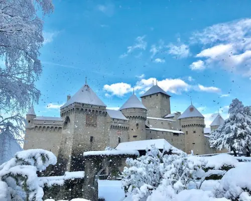 Chillon Castle in Switzerland covered in fresh snow during winter, under a bright blue sky, with surrounding trees and ground adorned in a magical white blanket.