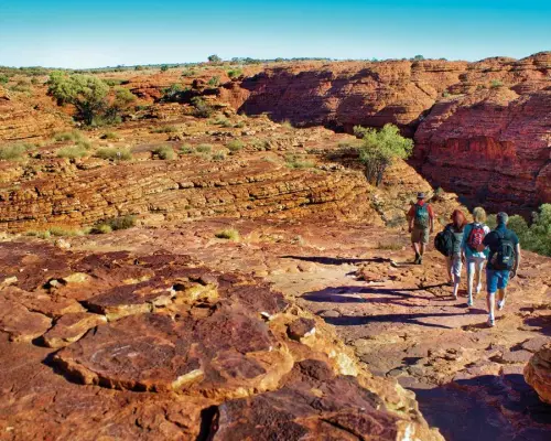 A group of backpack-clad tourists hiking on the Kings Canyon Rim Walk, admiring the magnificent red sandstone landscapes and vast outback panorama, experiencing thrilling adventure.