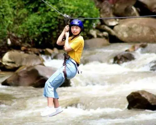 A female traveler enjoying the zipline adventure in Phuket, soaring over a rocky river and lush tropical rainforest with a joyful expression.