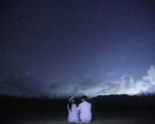 사이판 잔디밭에 함께 앉은 커플이 한국인 가이드와 함께 아름다운 별이 빛나는 밤하늘과 멀리 있는 구름을 감상하며 로맨틱한 스타라이트 투어를 경험하는 모습.