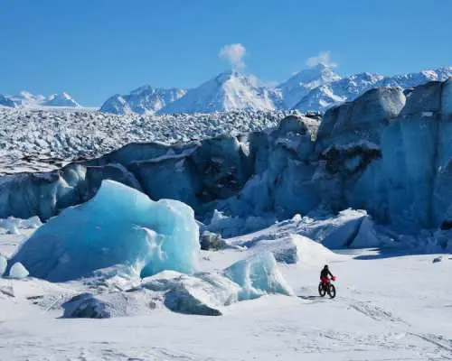 在阿拉斯加马塔努斯卡冰川的广阔雪地上，一位骑着雪地自行车的探险者，周围环绕着蓝色的巨型冰块与远方的雪山。