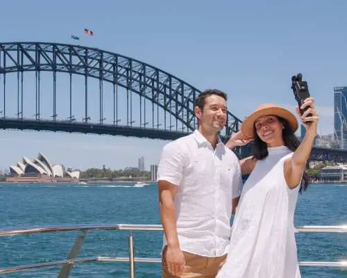 一對情侶在雪梨港渡輪上自拍，背景是宏偉的雪梨海港大橋和經典的雪梨歌劇院，輕鬆出示電子憑證即可搭乘。