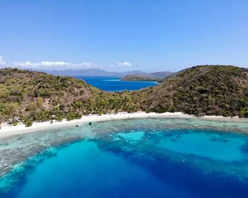 巴拉望科龍島嶼空拍圖，潔白粉狀沙灘弧線優美，廣闊湛藍海水中清晰可見的珊瑚礁，展現絕美潛水與浮潛天堂。
