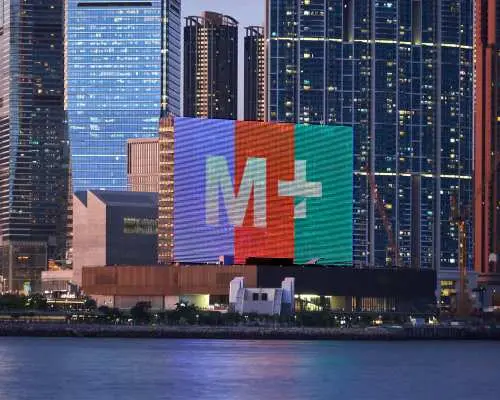 香港M+博物館迷人的夜間外觀，大型LED屏幕上M+標誌色彩斑斕，與香港絢麗的城市燈光及維港夜景交相輝映。