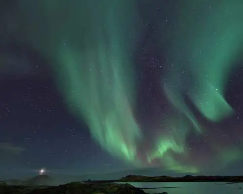 冰島夜空中絢爛的綠色極光如同瀑布般傾瀉而下，照亮了崎嶇的海岸線和遠方微亮的燈塔，展示極光船之旅的獨特視野。