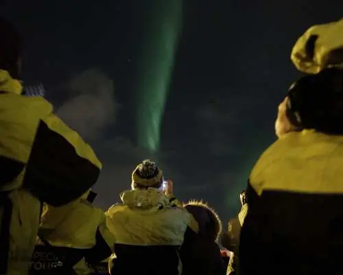 遊客穿著冬季大衣在冰島極光船上仰望夜空，欣賞一道道神秘的綠色極光，體驗海上追尋極光的獨特旅程。