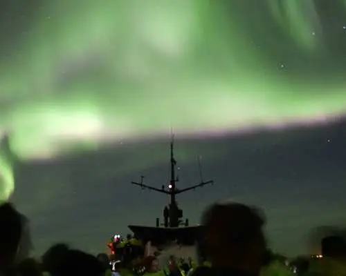 從冰島極光船甲板上望向遠方，遊客們興奮地仰望夜空中大片舞動的鮮綠色極光，享受難忘的海上奇觀。