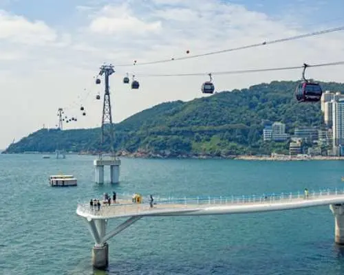 Busan Songdo Marine Cable Car cabins gliding over clear blue ocean waters, with coastal walkways and rocky outcrops below, and city buildings visible in the distance.