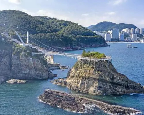 Busan Songdo Cloud Bridge connecting a small island to the mainland, spanning across the deep blue sea, surrounded by green hills and the city skyline.