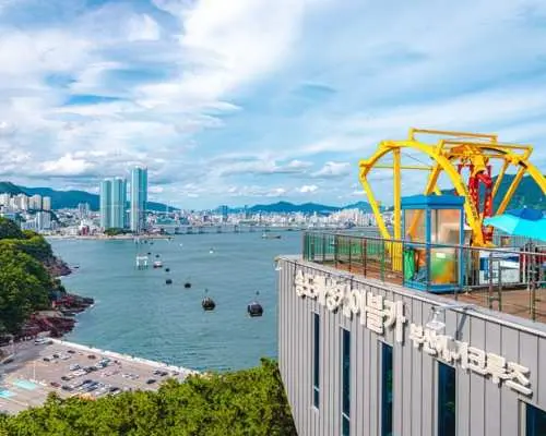 Busan Songdo Marine Cable Car station with vibrant yellow cabins and cable lines, offering panoramic views of the wide bay, city skyline, and distant mountains.