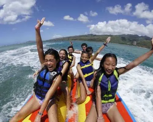 A joyful group of friends or family in life vests riding a vibrant banana boat across Oahu, Hawaii's blue waters, creating splashes and sharing an exciting group water adventure.