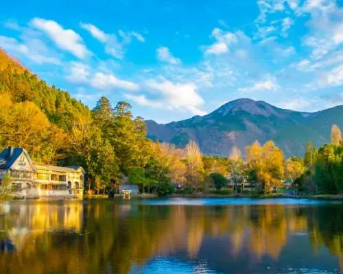 九州由布院金鱗湖畔，秋季楓葉染紅山林，湖面倒映建築與藍天白雲，如畫美景盡收眼底。