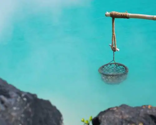 九州別府海地獄溫泉，水面呈碧藍色並冒著熱氣，吊掛煮食籃，展現當地溫泉文化與特色。