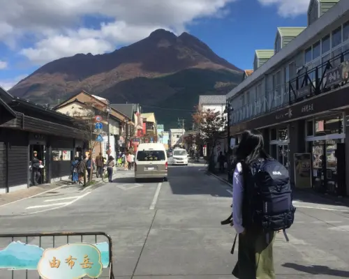 旅客背著背包在九州由布院街頭漫步，遠眺雄偉由布岳，感受小鎮獨特風情與自由旅行。