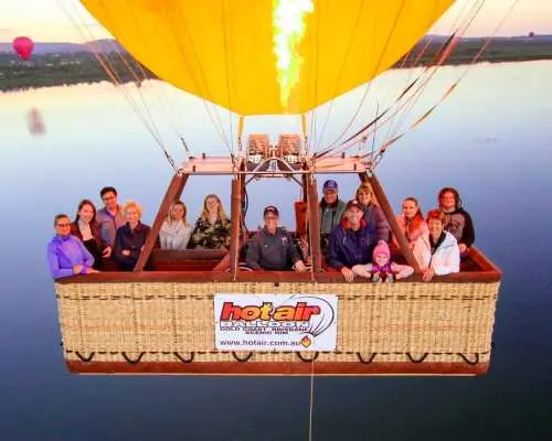 Passengers in a hot air balloon basket gaze out as flames ignite the canopy, overlooking Atherton Tablelands' serene lakes during this thrilling Port Douglas flight.