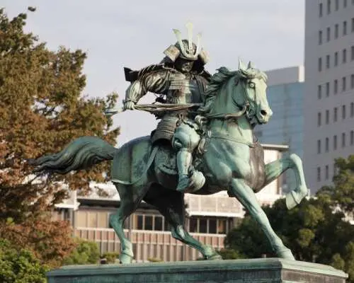 日本東京皇居外苑的楠木正成武士騎馬銅像，背景是現代建築，代表機場接送服務能便捷抵達東京的文化歷史景點。