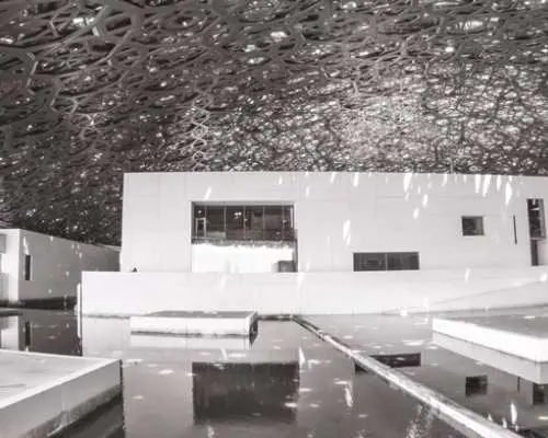 Inside Louvre Abu Dhabi, intricate woven dome structure overhead, with modern buildings beautifully reflected in the calm water below, creating unique light and shadow effects.