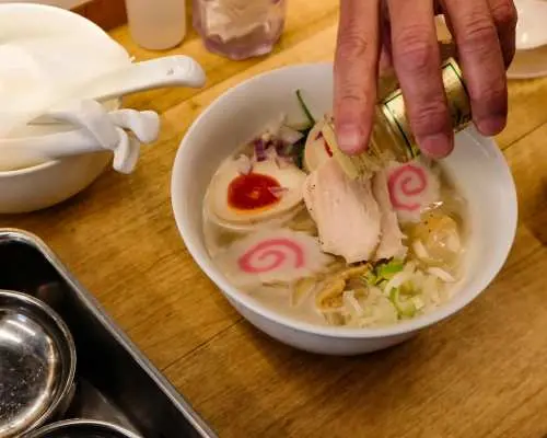 A hand garnishing a beautifully arranged bowl of professional-level ramen with chicken, egg, and narutomaki, showcasing the delicious result of an Osaka cooking class.