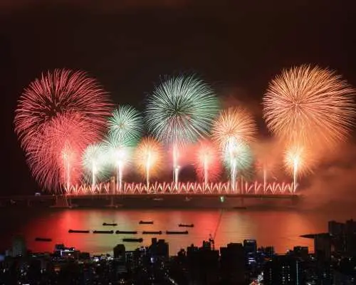 2025釜山煙火節在廣安大橋上空綻放紅色、綠色、橘色煙火，水中倒影與城市燈光交織成絕美畫面。