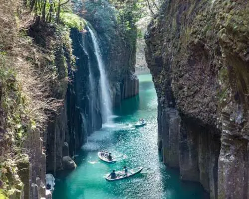 Visitors paddle boats in Takachiho Gorge, admiring the emerald green river and majestic Manai Waterfall, experiencing Miyazaki's volcanic natural wonders.