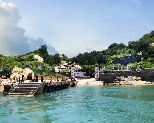 金門大膽島碼頭風光，遊客登島專用棧道與湛藍海水相映成趣，遠方綠意盎然的山丘與戰地建築隱約可見，適合半日探索。