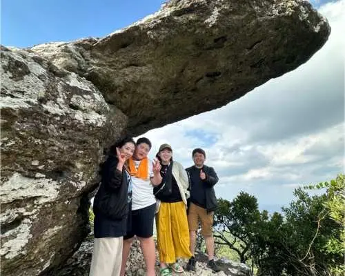 一群朋友在石垣島巨石頂下合影留念，背景是獨特的岩石地貌和翠綠樹林，結束電動三輪越野車健行後的歡樂時刻。