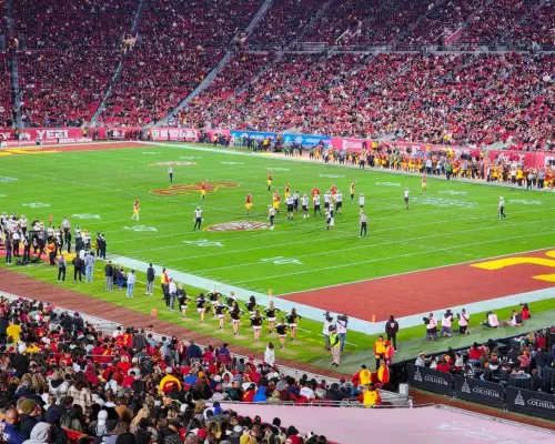 Trận bóng bầu dục USC Trojans đang diễn ra tại Sân vận động Tưởng niệm Los Angeles, với các cầu thủ và hoạt náo viên ở đường biên, cùng những người hâm mộ nhiệt tình mặc đồng phục đội lấp đầy khán đài, hòa mình vào văn hóa cổ vũ sôi động.