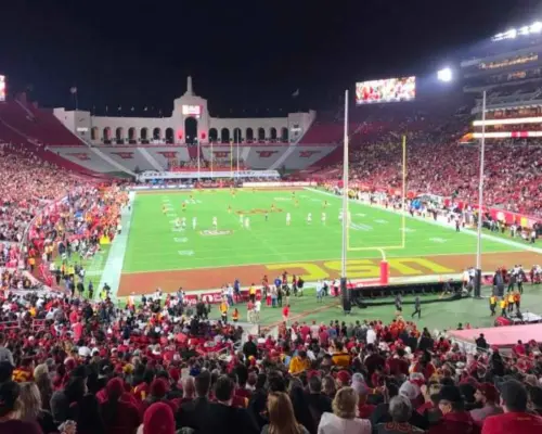 Toàn cảnh Sân vận động Tưởng niệm Los Angeles vào buổi tối trong trận bóng bầu dục USC Trojans, với kiến trúc lịch sử và biển người hâm mộ mặc đồ đỏ lấp đầy khán đài, trải nghiệm tinh thần bóng bầu dục đại học.