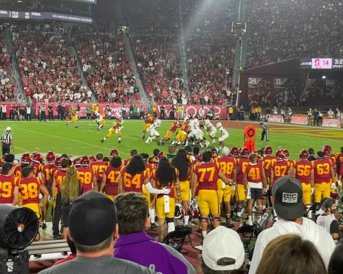 Góc nhìn cận cảnh từ khán đài ghi lại khoảnh khắc căng thẳng của trận bóng bầu dục USC Trojans, với các cầu thủ thi đấu trên sân và người hâm mộ cuồng nhiệt cổ vũ tại Sân vận động Tưởng niệm Los Angeles.