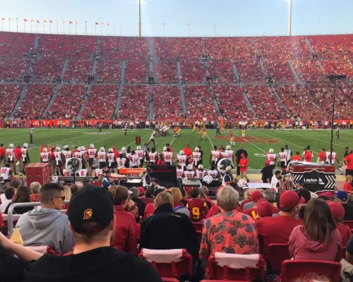 Một trận bóng bầu dục USC Trojans đầy kịch tính đang diễn ra tại Sân vận động Tưởng niệm Los Angeles, với người hâm mộ mặc màu áo đội trên khán đài, tận hưởng trải nghiệm xem trận đấu đầy phấn khích và đáng nhớ.