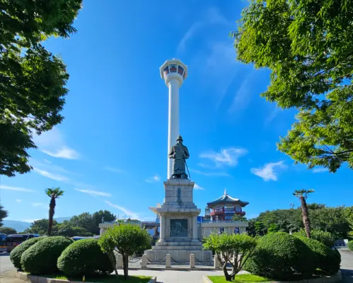 釜山龍頭山公園內，高聳的釜山塔與李舜臣將軍雕像在藍天綠樹環繞下，構成一幅和諧的地標景觀。
