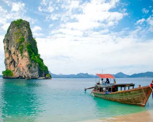 泰国甲米美丽的海岸线，一艘传统长尾船停泊在沙滩上，湛蓝海水与宏伟石灰岩喀斯特岛屿在晴空下，代表前往海岛的渡轮接送。