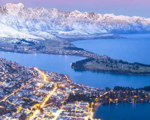 Queenstown dazzles at night, with city lights reflecting on Lake Wakatipu, framed by the dark, majestic snow-capped mountains under a twilight sky.