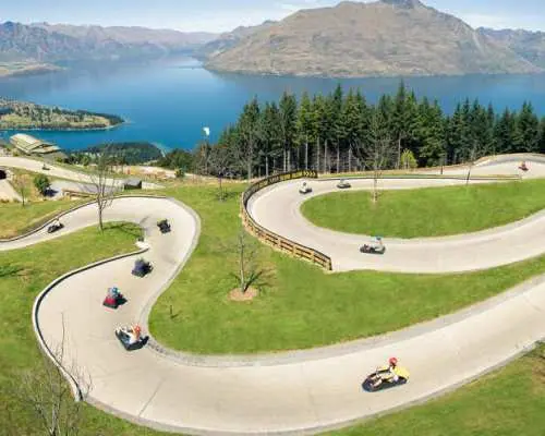 Adventurous guests race down a winding luge track at Skyline Queenstown, enjoying thrilling fun with stunning views of Lake Wakatipu and green hills.