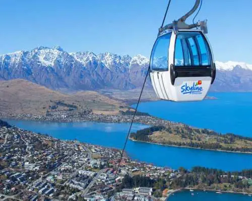 A blue Skyline gondola cabin ascends above Queenstown, offering aerial views of the vibrant town, serene Lake Wakatipu, and surrounding mountains.