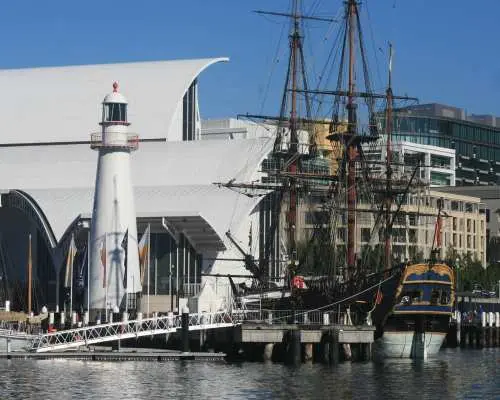 Waterfront view of the Australian National Maritime Museum in Sydney, featuring a white lighthouse and a majestic historic tall ship, possibly HMB Endeavour, docked, showcasing Australia's rich maritime past.
