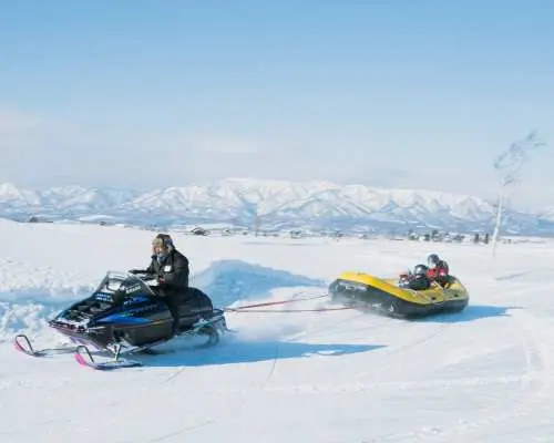 スノーモービルが興奮した乗客を乗せたインフレータブルラフトを広い雪原で牽引し、北海道でスリル満点の雪上ラフティングを提供しています。
