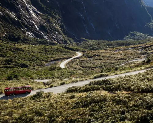 Red tour bus driving along a winding road through the vast, rugged wilderness with majestic mountains in the background, en route to Milford Sound from Queenstown.