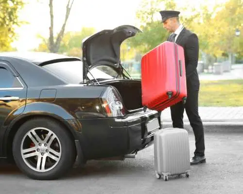 埃因霍温私人接送服务，专业司机身着制服，正协助旅客将红色大行李箱装入黑色豪华轿车后备箱。
