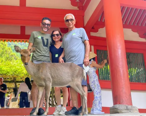 Tourists happily posing with a friendly Nara deer in Nara Park, with a vibrant vermilion Japanese shrine in the background, experiencing unique cultural interaction.