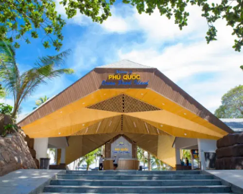푸꾸옥 명소 또는 호핑 투어 집합 장소의 환영 입구. 현대적인 디자인과 나무 액센트, 푸른 하늘 아래 무성한 열대 녹지가 특징입니다.