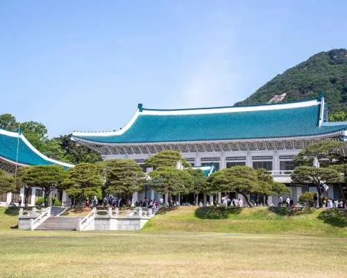 韩国首尔青瓦台，游客漫步于绿草如茵的广阔庭院，背景是蓝瓦屋顶与郁郁葱葱的山脉。