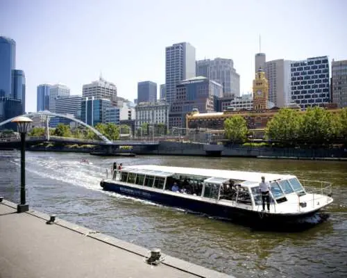 墨尔本雅拉河上舒适安全的观光游船，行驶于河畔，背景是城市地标建筑与绿树，船员在船头引导。