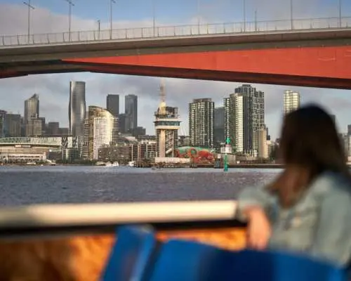 从雅拉河游船内部眺望墨尔本城市风光，高耸建筑在红色桥梁下若隐若现，游客舒适地欣赏河畔景色。